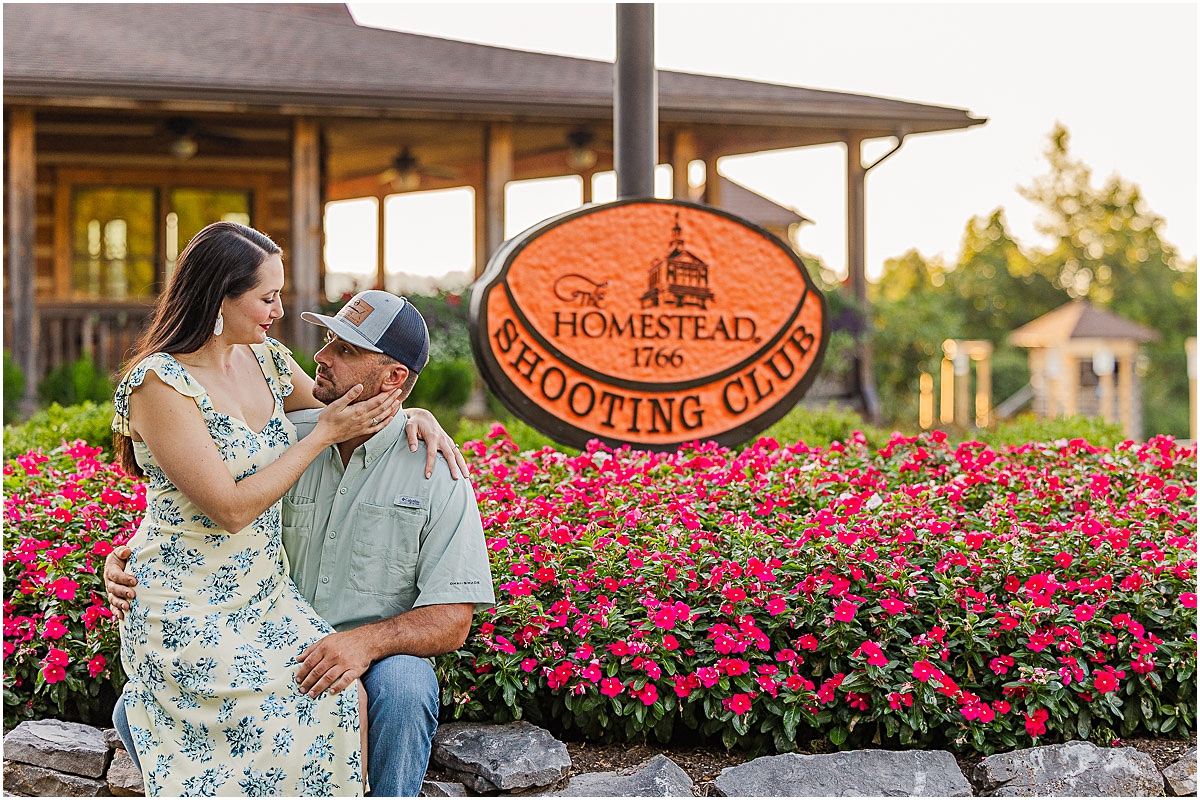 Mary sitting on Daniel's lap with a flower bed behind them and the sign for the Shooting Club during some engagement photography in Virginia
